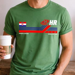 May include: A green short-sleeve t-shirt featuring a graphic with the Croatian flag, a soccer ball, and the text "CROATIA SOCCER" and "HR WORLD CUP 2026". The design includes horizontal blue and red stripes and three small stars.