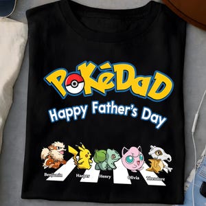 Può includere: T-shirt nera con la scritta "PokéDad Happy Father's Day" in giallo e blu. Sotto il testo ci sono personaggi dei cartoni animati e i nomi: Benjamin, Harper, Henry, Olivia e Theodore.
