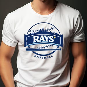 May include: White t-shirt featuring a baseball-themed design. The design includes the word "RAYS" in white letters on a blue banner, a baseball bat, and the word "BASEBALL". The design is set against a baseball and a cityscape.