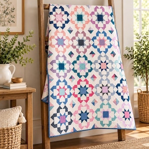 May include: A rectangular quilt featuring a repeating geometric star pattern in shades of pink, blue, purple, and teal on a white background. The quilt is draped over a wooden ladder, showcasing a multi-coloured patchwork design with intricate stitching.