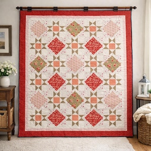 Op de afbeelding: Een grote, handgemaakte quilt met een witte achtergrond en een herhalend patroon van ruiten en sterren. De ruiten hebben bloemenprints in rood, roze en groen. De quilt is afgezet met een rode stippenstof en hangt aan een houten roede.