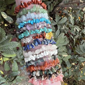 May include: A collection of gemstone chip bracelets in various colors, including red, green, blue, purple, and white. The bracelets are stacked on a pink cylindrical form, showcasing the variety of stones and textures. The background features green foliage.
