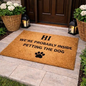 以下が含まれることがあります： 「HI WE'RE PROBABLY INSIDE PETTING THE DOG」の黒い文字と足跡がプリントされた長方形の天然繊維のドアマット。 茶色のドアの前に置かれています。