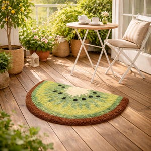 May include: A half-circle rug shaped like a kiwi fruit, with green, yellow, and brown colors. The rug is on a wooden deck, with a small table and chair in the background. The rug has a textured surface.