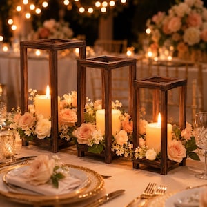 May include: Three rectangular dark brown wooden candle lanterns of varying heights, each containing a single lit pillar candle. The lanterns are surrounded by arrangements of light pink and white roses and small white filler flowers.