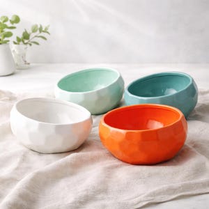May include: Four ceramic bowls in white, mint green, teal, and orange. Each bowl has a faceted exterior and a smooth, glossy interior. The bowls are arranged on a neutral-colored cloth, with a plant in the background.