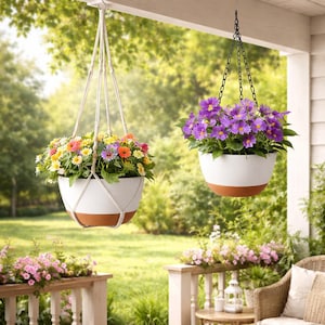 May include: Two hanging flower pots filled with colorful flowers. The pots are white with a brown base and are suspended by a macrame rope and a black chain. The background is a green, natural setting.