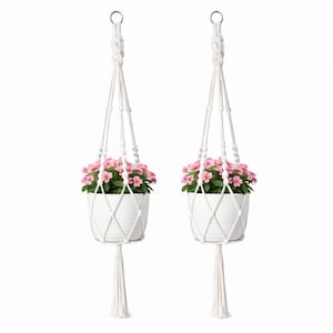 May include: Two white macrame plant hangers, each holding a white ceramic pot filled with pink flowers. The hangers have a woven design and a tassel at the bottom, suspended from a metal ring.