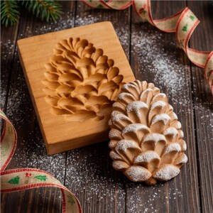 May include: A wooden pine cone cookie mold and a baked pine cone cookie dusted with white powder. The mold is rectangular with a carved pine cone design. A red and green ribbon and pine branches are in the background.