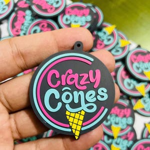 May include: A black, circular Crazy Cones charm featuring a pink and turquoise logo and a yellow ice cream cone graphic. The charm has a small hole for attachment. Similar charms are in the background.