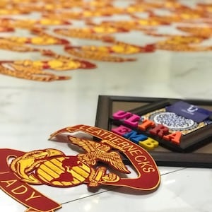 May include: Several red and gold embroidered patches with the words "Leathernecks" and "Lady" are visible. The patches feature an eagle, globe, and anchor design. A framed piece with colourful letters spelling "ULTRA EMB" is also present.