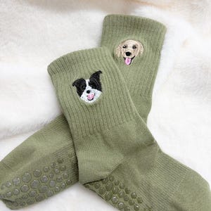 Può includere: Un paio di calzini verde oliva con facce di cani ricamate. Un calzino presenta un cane bianco e nero, l'altro un golden retriever. Le suole hanno delle prese antiscivolo.