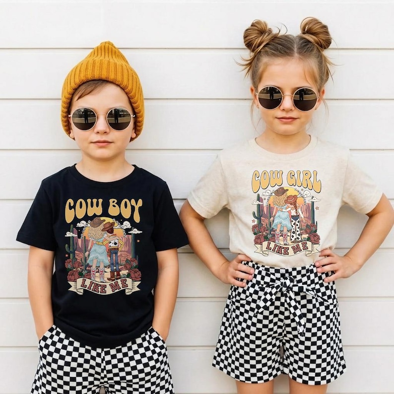 May include: A black t-shirt with a graphic print of a cowboy and the text COW BOY LIKE ME, and a cream t-shirt with a graphic print of a cowgirl and the text COW GIRL LIKE ME. Both shirts are paired with black and white checkered shorts.