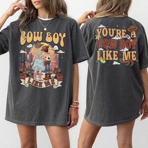May include: A charcoal gray short-sleeve t-shirt featuring a graphic print of two characters in a desert landscape. The front displays the text COW BOY LIKE ME. The back features the text YOU'RE A COW BOY LIKE ME with icons of a hat, rocket, and ball.