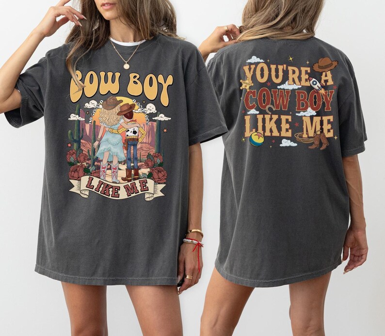 May include: A charcoal gray short-sleeve t-shirt featuring a graphic print of two characters in a desert landscape. The front displays the text COW BOY LIKE ME. The back features the text YOU'RE A COW BOY LIKE ME with icons of a hat, rocket, and ball.
