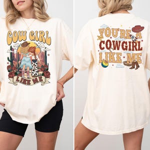 May include: An off-white short-sleeve t-shirt featuring a graphic print. The front displays two figures in a desert landscape with the text COW GIRL LIKE ME. The back features western-themed icons and the text YOU'RE A COWGIRL LIKE ME.