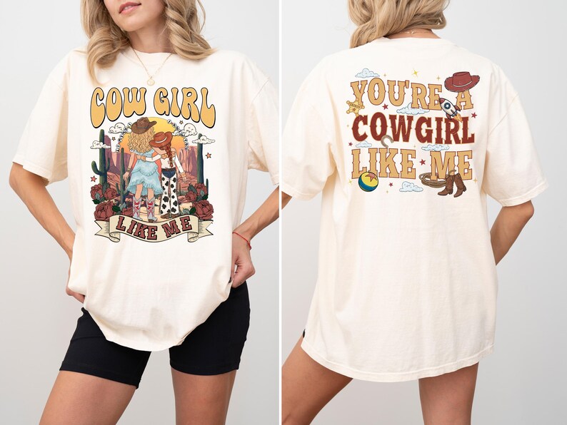May include: An off-white short-sleeve t-shirt featuring a graphic print. The front displays two figures in a desert landscape with the text COW GIRL LIKE ME. The back features western-themed icons and the text YOU'RE A COWGIRL LIKE ME.