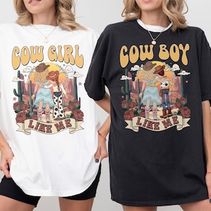 May include: A white t-shirt and a black t-shirt, each featuring a graphic print of a desert scene with cacti, roses, and two figures. The white shirt says COW GIRL LIKE ME, and the black shirt says COW BOY LIKE ME, both in a retro, curved font.
