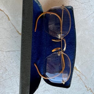 May include: Vintage eyeglasses with gold-toned frames and clear lenses rest inside a dark blue velvet-lined case. The case is black with a textured exterior. The glasses have a classic, rimless design.