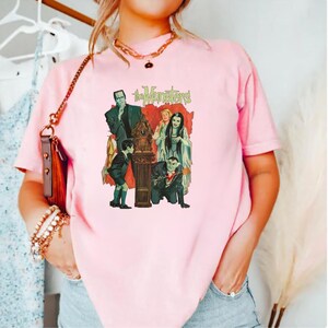 Peut inclure: T-shirt rose clair avec un graphisme vintage de la famille Munster. Le design comprend les mots "The Munsters" en vert, avec les membres de la famille dans leurs tenues classiques. Un design de style r&eacute;tro.