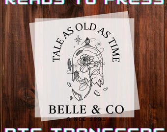 May include: A DTF transfer sheet with the text "READY TO PRESS" and "DTF TRANSFERS". The design features a rose inside a broken glass dome with the text "TALE AS OLD AS TIME" and "BELLE & CO SINCE 1991". The image is on a wooden background.
