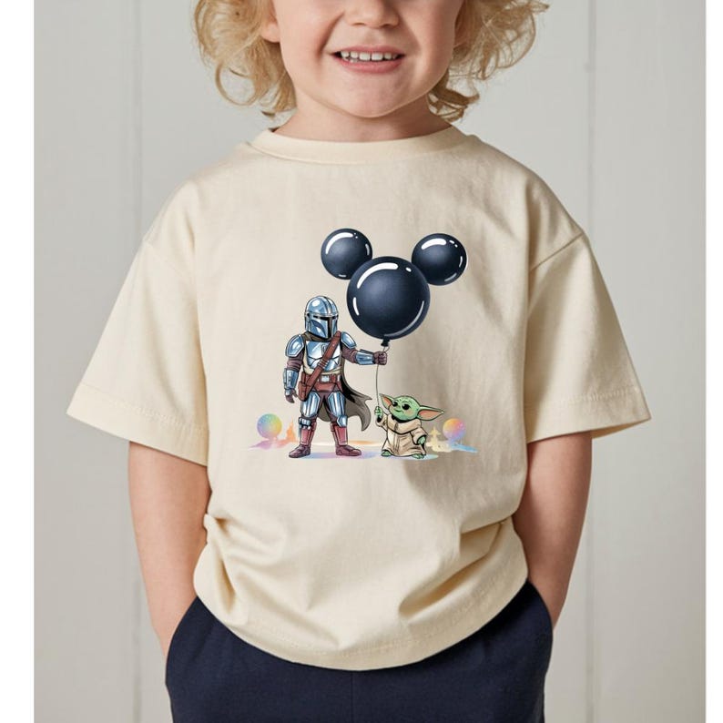May include: Cream-colored t-shirt featuring a cartoon illustration of The Mandalorian and Grogu. The Mandalorian is holding a black balloon in the shape of Mickey Mouse ears. Grogu is standing next to him. Colorful orbs are in the background.