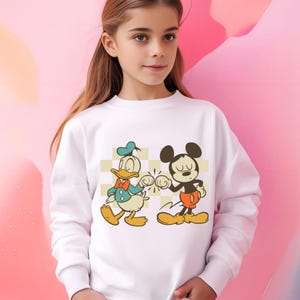 May include: White sweatshirt featuring a graphic of Donald Duck and Mickey Mouse bumping fists. Donald Duck is wearing a blue hat and jacket with a red bow tie. Mickey Mouse is wearing red shorts and yellow shoes. The background is a checkered pattern.