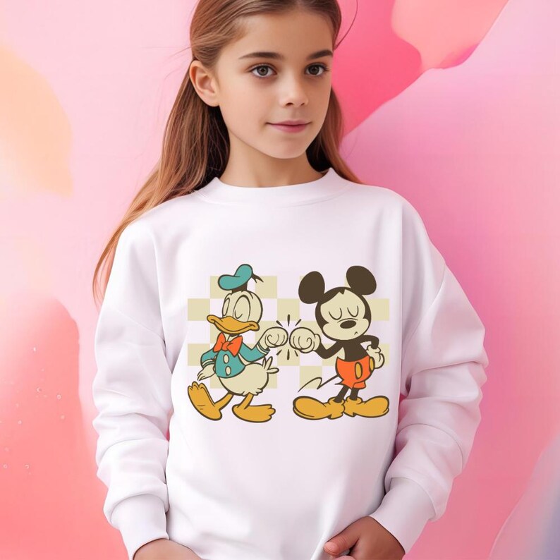 May include: White sweatshirt featuring a graphic of Donald Duck and Mickey Mouse bumping fists. Donald Duck is wearing a blue hat and jacket with a red bow tie. Mickey Mouse is wearing red shorts and yellow shoes. The background is a checkered pattern.