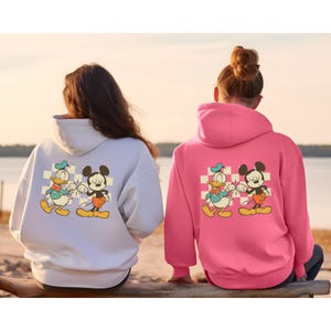 May include: White and pink hoodies featuring a cartoon graphic of Mickey Mouse and Donald Duck. The cartoon characters are set against a checkered background. The hoodies have a hood and long sleeves.