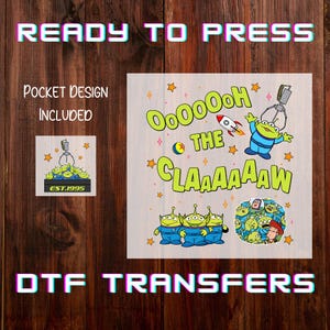 May include: DTF transfers featuring a design with the text "OooOoooh the Claaaaaaw" and cartoon aliens. The image includes a pocket design and the text "Ready to Press". The design is on a wooden background.