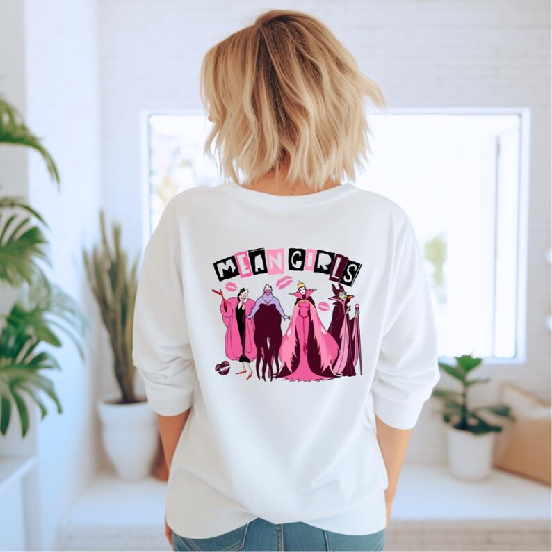 May include: White long-sleeved sweatshirt with a graphic on the back. The graphic features the text "MEAN GIRLS" in pink and black block letters, with cartoon illustrations of characters in pink and purple outfits beneath.