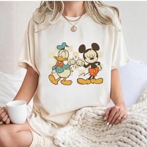 May include: Cream-colored t-shirt featuring a cartoon graphic of Donald Duck and Mickey Mouse bumping fists. Donald Duck is wearing a blue sailor suit and Mickey Mouse is in red shorts. The background has a yellow and white checkered pattern.