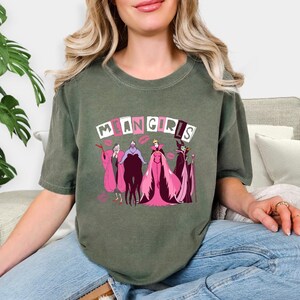 May include: An olive green t-shirt with the words "MEAN GIRLS" above a cartoon graphic of villains in pink and purple. The design includes lipstick kiss marks. The shirt is a casual style.