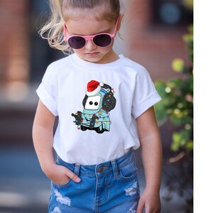 A young girl wearing a white t-shirt with a cartoon character and sunglasses, standing outdoors.