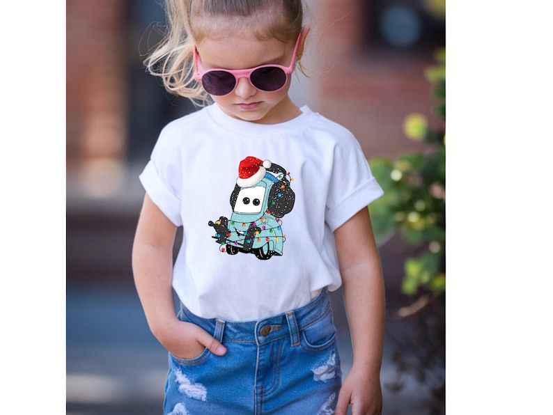 A young girl wearing a white t-shirt with a cartoon character and sunglasses, standing outdoors.
