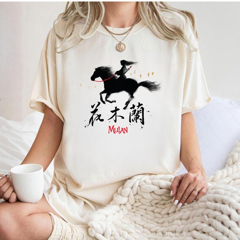 Pode incluir: Camiseta creme com uma silhueta preta de uma pessoa montada a cavalo, com o texto "Mulan" em vermelho e caracteres chineses. O desenho est&aacute; centrado na frente da camiseta.
