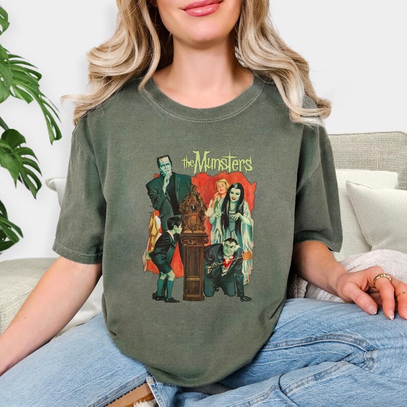 Peut inclure: T-shirt vert olive avec un motif de la famille Munster et le texte "the Munsters". Le motif repr&eacute;sente les personnages de la s&eacute;rie t&eacute;l&eacute;vis&eacute;e classique. Le t-shirt est de style d&eacute;contract&eacute; et confortable.