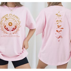 Op de afbeelding: Lichtroze T-shirt met een opdruk op de voorkant. De opdruk toont de tekst "IRON FLAME TURN IT DOWN" in goud en rood, omgeven door een cirkelvormig ontwerp. De achterkant heeft een reeks gestileerde draken in goud- en roodtinten.