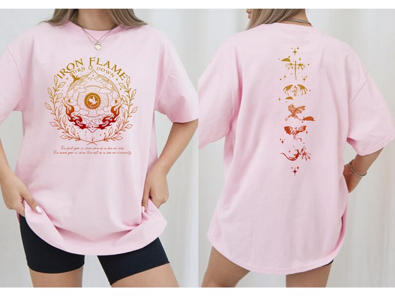 Op de afbeelding: Lichtroze T-shirt met een opdruk op de voorkant. De opdruk toont de tekst "IRON FLAME TURN IT DOWN" in goud en rood, omgeven door een cirkelvormig ontwerp. De achterkant heeft een reeks gestileerde draken in goud- en roodtinten.