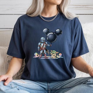 May include: Navy blue t-shirt featuring a graphic of The Mandalorian and Grogu holding a Mickey Mouse-shaped balloon. The illustration includes colorful accents. The shirt is a comfortable, casual wear item.