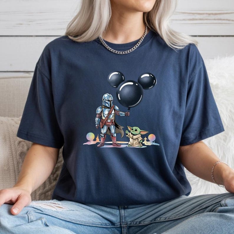 May include: Navy blue t-shirt featuring a graphic of The Mandalorian and Grogu holding a Mickey Mouse-shaped balloon. The illustration includes colorful accents. The shirt is a comfortable, casual wear item.