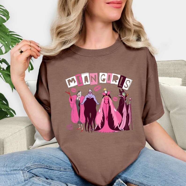 May include: A brown t-shirt with the words "MEAN GIRLS" above a cartoon graphic of villains in pink and purple. The design includes lipstick kiss marks. The t-shirt is a casual style.