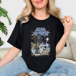 May include: Black t-shirt with a Star Wars graphic. The design features Darth Vader, other characters, and the Star Wars logo in blue and white. The shirt has a classic crew neck.