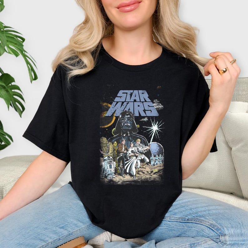 May include: Black t-shirt with a Star Wars graphic. The design features Darth Vader, other characters, and the Star Wars logo in blue and white. The shirt has a classic crew neck.