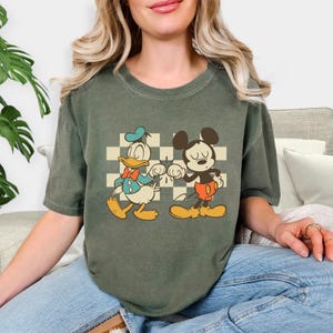 May include: Olive green t-shirt featuring a vintage-style graphic of Donald Duck and Mickey Mouse bumping fists in front of a checkered background. The cartoon characters are in their classic outfits.