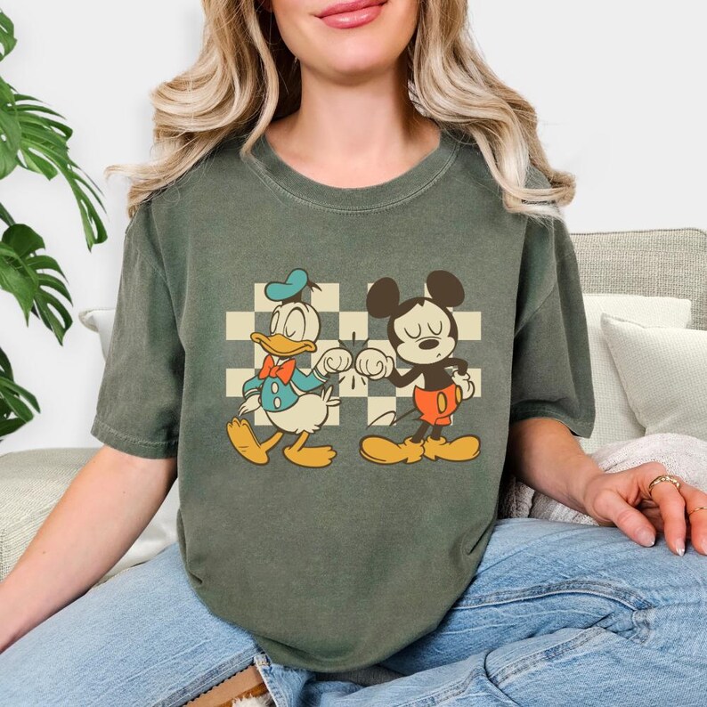 May include: Olive green t-shirt featuring a vintage-style graphic of Donald Duck and Mickey Mouse bumping fists in front of a checkered background. The cartoon characters are in their classic outfits.