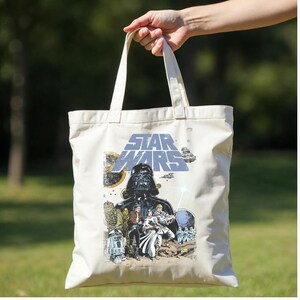 May include: Cream-coloured tote bag featuring a Star Wars graphic. The design includes Darth Vader, other characters, and the Star Wars logo in blue. The bag has two white handles and is held up by a hand.