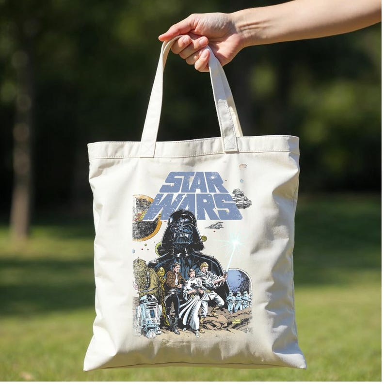 May include: Cream-coloured tote bag featuring a Star Wars graphic. The design includes Darth Vader, other characters, and the Star Wars logo in blue. The bag has two white handles and is held up by a hand.
