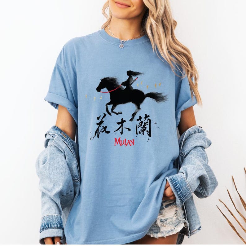 Pode incluir: T-shirt azul claro com um desenho preto de uma pessoa a cavalo, acompanhado de caracteres chineses e a palavra "MULAN" em vermelho. O desenho &eacute; impresso na frente da t-shirt.