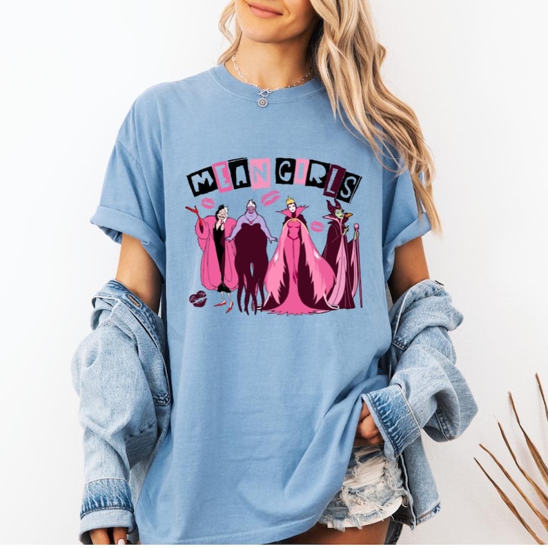 May include: Pale blue t-shirt with the text "MEAN GIRLS" and a graphic of cartoon villains in pink and purple. The design includes characters in robes and dresses. The shirt has short sleeves and a loose fit.